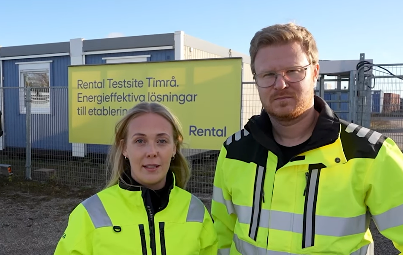 Två personer, en kvinna och en man, står utomhus framför en skylt och en byggmodul. Båda bär ljusgula varseljackor. Skylten i bakgrunden är gul och har texten "Rental Testsite Timrå. Energieffektiva lösningar till etableringen.